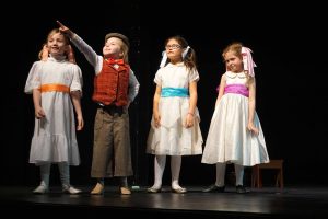 Young Kids On Stage actors perform on stage in costume, with one child pointing upward during a children’s theater scene.
